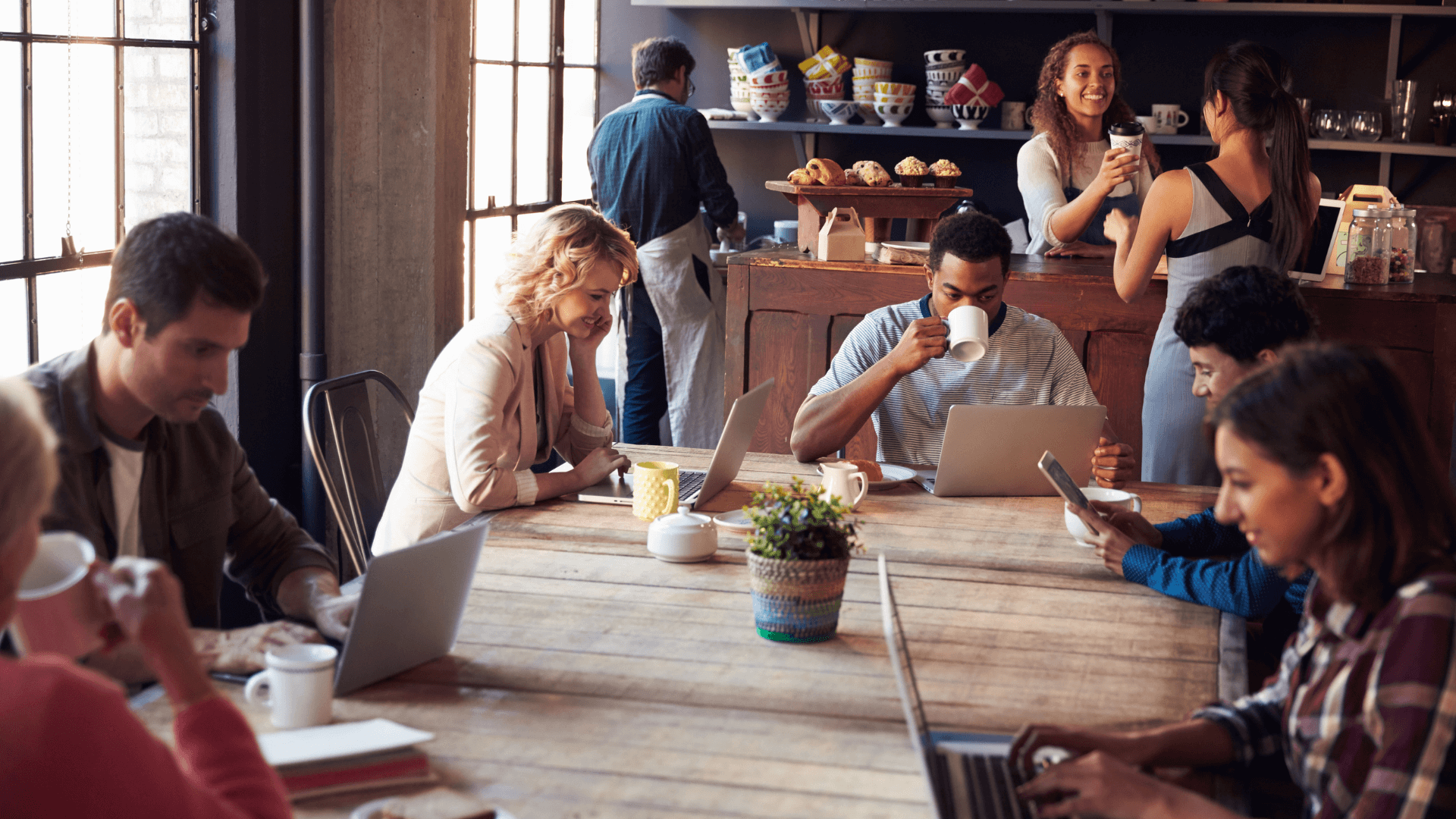 Menschen sitzen an einem langen Holztisch in einem Café und arbeiten an Laptops und Tablets, während sie Kaffee trinken. Im Hintergrund bereiten zwei Baristas Getränke zu, und ein Mann holt sich etwas von der Theke. Die Atmosphäre ist lebhaft und produktiv.